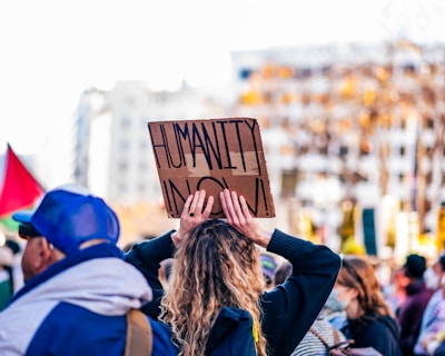 A crowd of people is gathered at an outdoor event, with one person holding a cardboard sign that reads 'HUMANITY NOW'. The scene includes various individuals wearing casual clothing, some with hats and masks. The background features urban elements and blurred foliage, suggesting a public demonstration or protest.