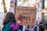A child is holding a cardboard sign with the message 'Fund Childcare Not Wars' written on it, featuring colorful scribbles and drawings. The background shows a city street with blurred people and lights, suggesting a protest or demonstration setting.