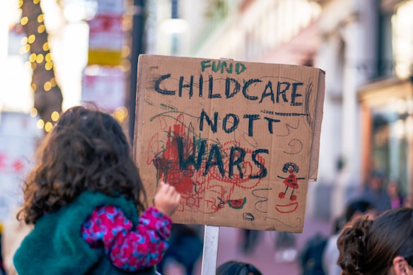 A child is holding a cardboard sign with the message 'Fund Childcare Not Wars' written on it, featuring colorful scribbles and drawings. The background shows a city street with blurred people and lights, suggesting a protest or demonstration setting.