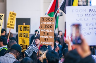 A crowd of people is gathered with various protest signs. Some signs bear messages about children, women, medical personnel, and journalists, while another highlights the phrase 'No Xmas as usual in a genocide'. People are clustered closely, some with hats or scarves, and flags are present in the background, contributing to the demonstration's atmosphere.