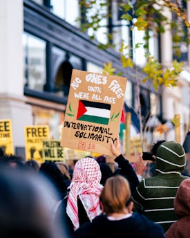 A group of people stand in a protest, holding signs with messages of solidarity and freedom. One prominent sign features the Palestinian flag with the words 'No one's free until we're all free' and 'International solidarity.' Participants wear clothing with cultural patterns and some are using phones to capture the event.