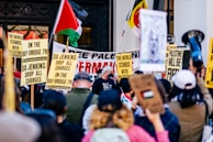 A large group of people gather at a protest holding various signs and flags supporting Palestine. The signs include messages like 'The World Stands with Palestine' and 'DA Jenkins: Drop All Charges on the Bay Bridge 78.' A Palestinian flag is prominently displayed among the crowd.