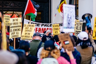A large group of people gather at a protest holding various signs and flags supporting Palestine. The signs include messages like 'The World Stands with Palestine' and 'DA Jenkins: Drop All Charges on the Bay Bridge 78.' A Palestinian flag is prominently displayed among the crowd.