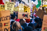 A crowd of people gathers for a protest or demonstration, holding various signs and banners with political messages. Prominent words include 'Solidarity', 'Palestine', and 'Genocide'. One sign features a drawing of a black dog with the word 'Forever' written below it. The scene is lively and packed with individuals actively participating.