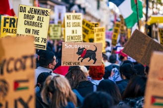 A crowd of people gathers for a protest or demonstration, holding various signs and banners with political messages. Prominent words include 'Solidarity', 'Palestine', and 'Genocide'. One sign features a drawing of a black dog with the word 'Forever' written below it. The scene is lively and packed with individuals actively participating.