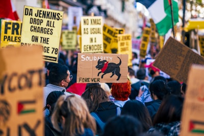 A crowd of people gathers for a protest or demonstration, holding various signs and banners with political messages. Prominent words include 'Solidarity', 'Palestine', and 'Genocide'. One sign features a drawing of a black dog with the word 'Forever' written below it. The scene is lively and packed with individuals actively participating.