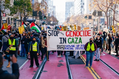 A large group of people is gathered on a city street, holding various signs and flags. The main sign in the center reads, 'Stop the genocide in Gaza,' with additional text emphasizing racial justice. Some participants wear yellow safety vests and face masks. The scene is vibrant with activity, as more signs are visible in the background, promoting messages in support of Palestinians.
