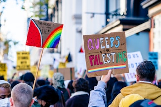 A crowd of people are gathered, holding up protest signs. One sign reads 'Pinkwashing is a damn lie!' with a rainbow and watermelon design, and another says 'Queers for a free Palestine' with colorful text on a cardboard background. People are dressed in casual clothing, some wearing jackets.