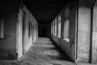 A black-and-white photograph of an empty stone corridor featuring a series of arched windows on one side and a stone wall on the other. The corridor is illuminated by soft light coming through the windows, casting shadows on the stone tile floor. The ceiling is made of dark wood, adding a historical and architectural atmosphere to the scene.