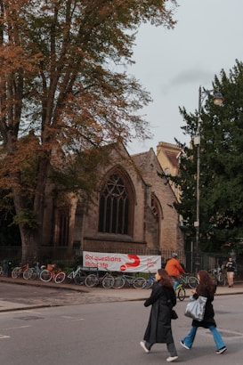 A historic church building with large windows and a fence in front, surrounded by trees. Several bicycles are parked along the sidewalk, and a banner with text hangs on the fence. People are walking and cycling on the street, with one person wearing an orange jacket riding a bicycle.