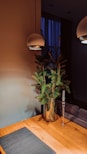 A minimalist interior design features a wooden table with a dark placemat, a glass vase holding green pine branches, a tall glass candle holder without a candle, and two modern overhead pendant lights. The background includes a dim, softly lit room with a blue curtain covering a window.
