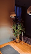 A minimalist interior design features a wooden table with a dark placemat, a glass vase holding green pine branches, a tall glass candle holder without a candle, and two modern overhead pendant lights. The background includes a dim, softly lit room with a blue curtain covering a window.