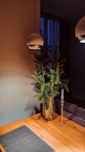 A minimalist interior design features a wooden table with a dark placemat, a glass vase holding green pine branches, a tall glass candle holder without a candle, and two modern overhead pendant lights. The background includes a dim, softly lit room with a blue curtain covering a window.