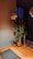 A minimalist interior design features a wooden table with a dark placemat, a glass vase holding green pine branches, a tall glass candle holder without a candle, and two modern overhead pendant lights. The background includes a dim, softly lit room with a blue curtain covering a window.
