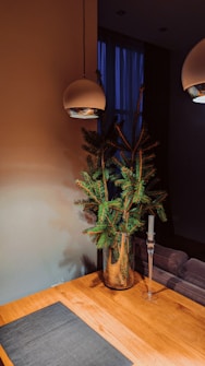 A minimalist interior design features a wooden table with a dark placemat, a glass vase holding green pine branches, a tall glass candle holder without a candle, and two modern overhead pendant lights. The background includes a dim, softly lit room with a blue curtain covering a window.