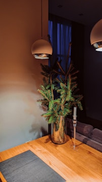 A minimalist interior design features a wooden table with a dark placemat, a glass vase holding green pine branches, a tall glass candle holder without a candle, and two modern overhead pendant lights. The background includes a dim, softly lit room with a blue curtain covering a window.