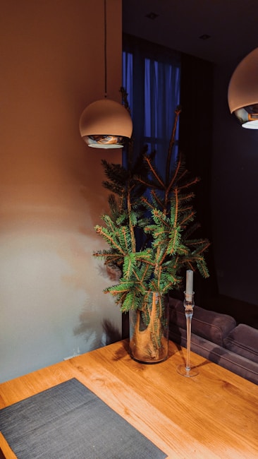 A minimalist interior design features a wooden table with a dark placemat, a glass vase holding green pine branches, a tall glass candle holder without a candle, and two modern overhead pendant lights. The background includes a dim, softly lit room with a blue curtain covering a window.