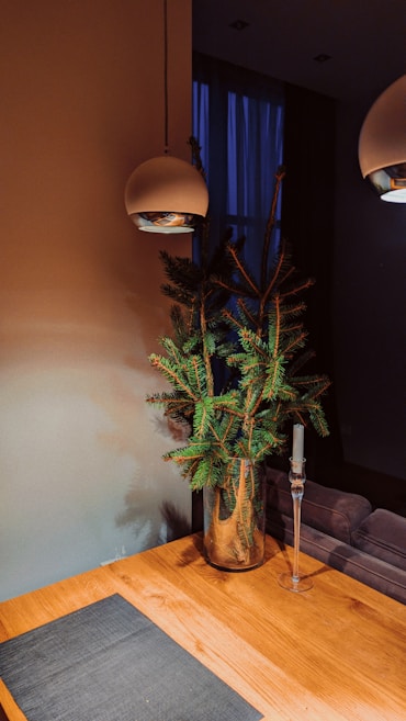 A minimalist interior design features a wooden table with a dark placemat, a glass vase holding green pine branches, a tall glass candle holder without a candle, and two modern overhead pendant lights. The background includes a dim, softly lit room with a blue curtain covering a window.