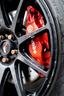 A close-up shot of a red Brembo brake caliper mounted on a dark alloy wheel of a vehicle. The metallic surface of the wheel complements the vibrant red of the caliper, which bears the white Brembo logo. The background shows part of the brake disc.