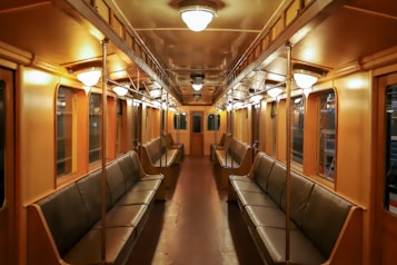 A vintage-style train car interior with wooden paneling and leather seats. The car is empty, showcasing evenly spaced lighting and a long aisle between two rows of benches. The ambient lighting lends a warm and nostalgic feel, with reflections visible in the side windows.