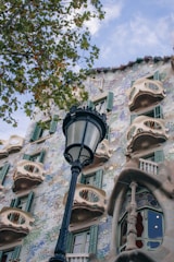 A decorative building facade with ornate elements and curved balconies is complemented by a black streetlamp in the foreground. The building features green shuttered windows and a mixture of pastel colors in the exterior design. Leafy branches from a tree partly frame the scene on the left.