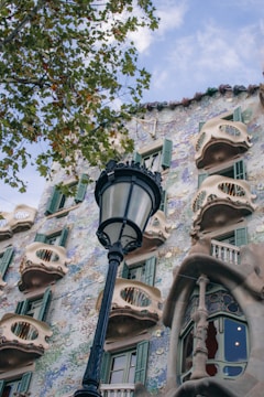 A decorative building facade with ornate elements and curved balconies is complemented by a black streetlamp in the foreground. The building features green shuttered windows and a mixture of pastel colors in the exterior design. Leafy branches from a tree partly frame the scene on the left.