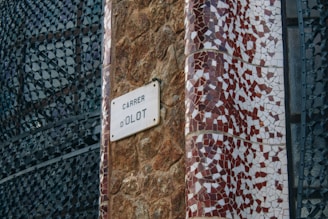 A street sign reading 'Carrer d'Olot' is affixed to a textured stone wall. The area next to the sign is adorned with an intricate mosaic pattern featuring red and white tiles. The background includes an artistic metal lattice structure with abstract patterns.