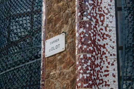 A street sign reading 'Carrer d'Olot' is affixed to a textured stone wall. The area next to the sign is adorned with an intricate mosaic pattern featuring red and white tiles. The background includes an artistic metal lattice structure with abstract patterns.