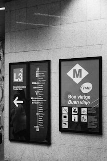 Two metro informational signs are mounted on a tiled wall. The left sign displays the L3 metro line with a list of stops and a directional arrow. The right sign has metro-related symbols and messages in Catalan and Spanish, including 'Bon viatge' and 'Buen viaje'.