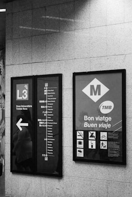Two metro informational signs are mounted on a tiled wall. The left sign displays the L3 metro line with a list of stops and a directional arrow. The right sign has metro-related symbols and messages in Catalan and Spanish, including 'Bon viatge' and 'Buen viaje'.