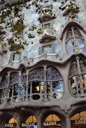 An ornate building facade featuring highly intricate architectural details, with sculptural elements and flowing organic forms. The facade includes unique windows and mosaic-like patterns, complemented by vibrant greenery.