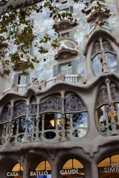 An ornate building facade featuring highly intricate architectural details, with sculptural elements and flowing organic forms. The facade includes unique windows and mosaic-like patterns, complemented by vibrant greenery.