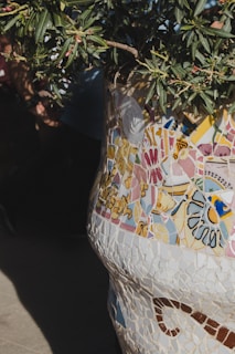 A detailed and colorful mosaic art piece with various patterns and colors, primarily composed of fragments with floral designs and abstract shapes. At the top, there is a plant with green leaves extending out.