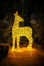 A luminous deer-shaped decoration made of interwoven golden lights stands in a natural setting, surrounded by trees. The glowing figure emits a warm and inviting light, illuminating the immediate snowy surroundings.