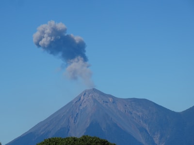 A mountain with a peak releasing a plume of smoke into the clear blue sky, indicating volcanic activity. The landscape is dominated by the steep slopes and rugged terrain of the volcano.