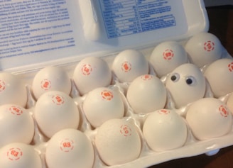 An egg carton holds a dozen white eggs, each marked with a red stamp. Among them, one egg stands out with googly eyes attached, adding a playful element to the scene.
