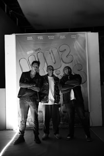 Three men are standing in front of a wall with their arms crossed. The background features a banner with text and graphics. The lighting is dim, with a spotlight casting shadows.
