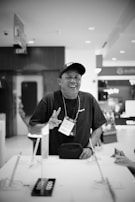 A person wearing a cap and a lanyard with a badge labeled 'CREW' is standing at a counter. They are smiling and gesturing with their hand. The setting appears to be indoors, with various objects on the counter and commercial signs in the background.