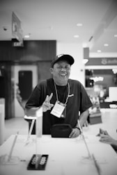 A person wearing a cap and a lanyard with a badge labeled 'CREW' is standing at a counter. They are smiling and gesturing with their hand. The setting appears to be indoors, with various objects on the counter and commercial signs in the background.