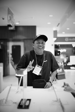 A person wearing a cap and a lanyard with a badge labeled 'CREW' is standing at a counter. They are smiling and gesturing with their hand. The setting appears to be indoors, with various objects on the counter and commercial signs in the background.