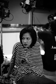 A woman with short hair and a striped shirt sits with a thoughtful expression in what appears to be a studio setting. Lighting equipment and a camera are visible in the background, indicating a possible filming or photography session is taking place. Other people are partially visible, engaged in activities out of focus.
