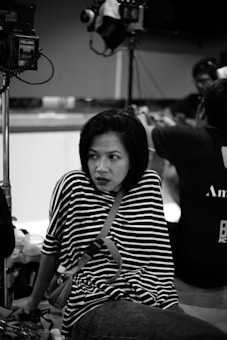 A woman with short hair and a striped shirt sits with a thoughtful expression in what appears to be a studio setting. Lighting equipment and a camera are visible in the background, indicating a possible filming or photography session is taking place. Other people are partially visible, engaged in activities out of focus.