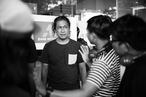 A group of people gathered outside a bank. The central figure, wearing a dark shirt with a checkered pocket, is listening intently to another person gesturing with a hand outstretched. The setting appears to be a casual meeting or discussion.