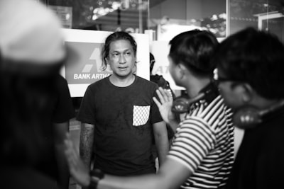 A group of people gathered outside a bank. The central figure, wearing a dark shirt with a checkered pocket, is listening intently to another person gesturing with a hand outstretched. The setting appears to be a casual meeting or discussion.