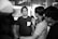 A group of people gathered outside a bank. The central figure, wearing a dark shirt with a checkered pocket, is listening intently to another person gesturing with a hand outstretched. The setting appears to be a casual meeting or discussion.