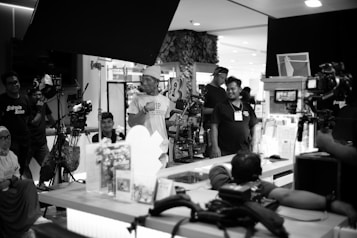 A group of people working on a film set with various equipment such as cameras and lights. Some individuals are engaged in conversation, while others focus on operating equipment. The setting appears indoors, and the mood is collaborative.