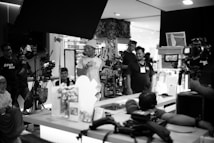 A group of people working on a film set with various equipment such as cameras and lights. Some individuals are engaged in conversation, while others focus on operating equipment. The setting appears indoors, and the mood is collaborative.