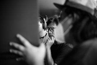 A black and white photo capturing a focused group of people. The central figure, wearing glasses, appears to be in deep thought, resting their chin on their hand. Two other individuals are visible, one in the foreground wearing a cap, and another beside the central figure wearing headphones.