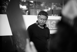 A person wearing a baseball cap backwards and headphones around the neck appears to be in thought or concentration. The background shows a blurry interior setting, possibly an office or studio environment, with some signage visible.