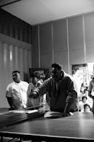 A group of people gather around a ping pong table indoors, with one person appearing to be in action, possibly making a move in a game. The scene is captured in black and white, and the background features a banner and an audience observing the activity.